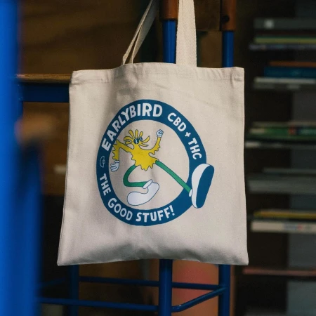 Canvas tote bag featuring 'Earlybird CBD + THC' logo with a cartoon bird character, hanging on a blue metal rack. Eco-friendly branding.