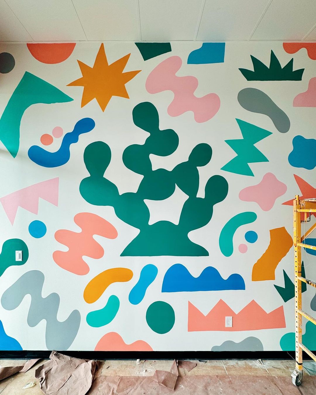 Abstract mural art featuring a green cactus and colorful geometric shapes on a white wall inside a building with natural light.