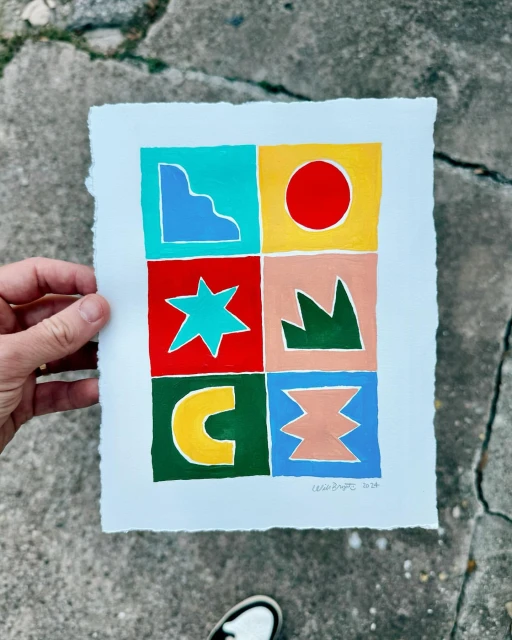 Abstract geometric gouache painting with bright colors and shapes, held in a person's hand against a concrete background.
