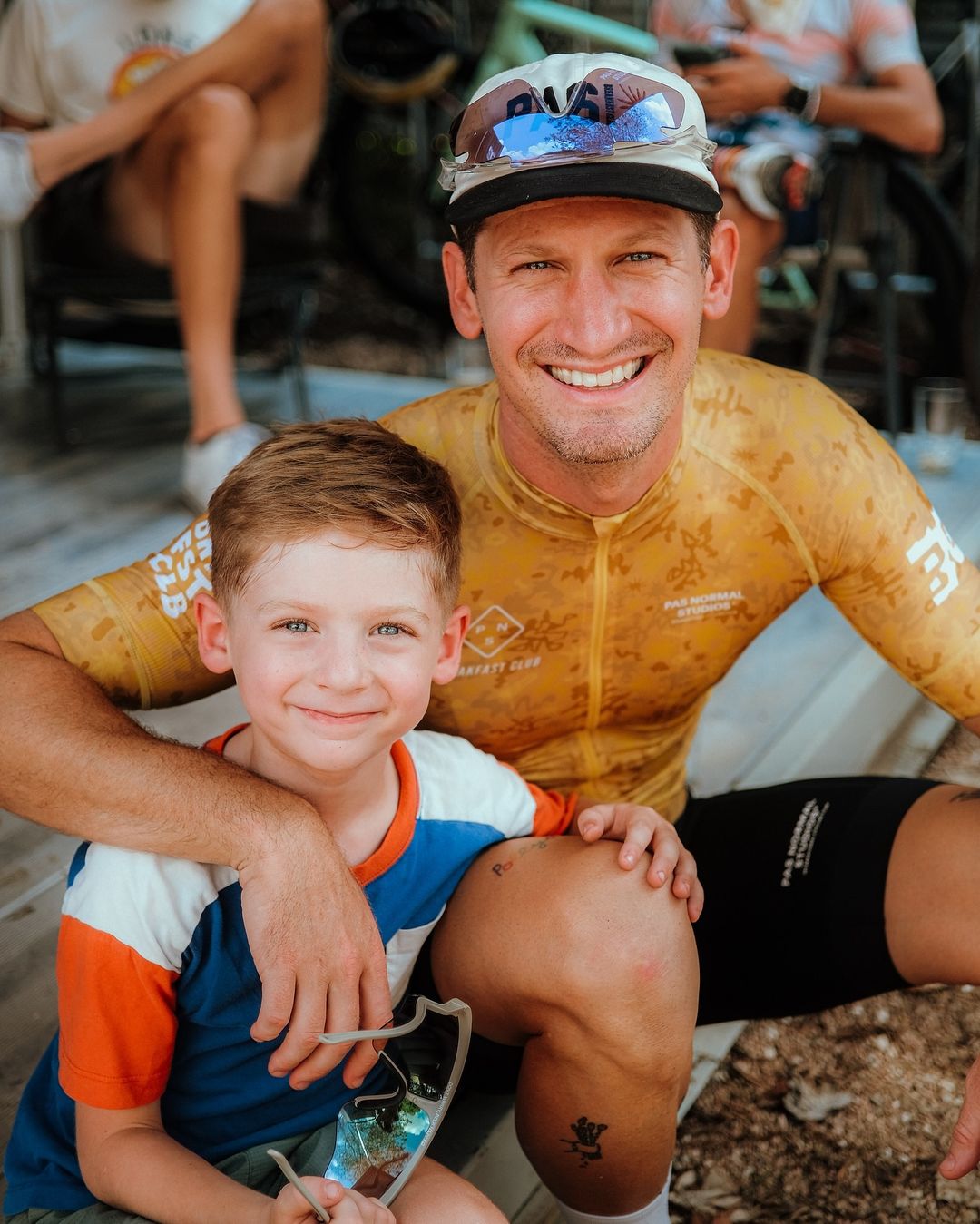 Smiling father and son posing together in a close shot, with the father wearing cycling gear and the child holding sunglasses.