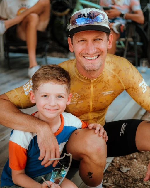Smiling father and son posing together in a close shot, with the father wearing cycling gear and the child holding sunglasses.