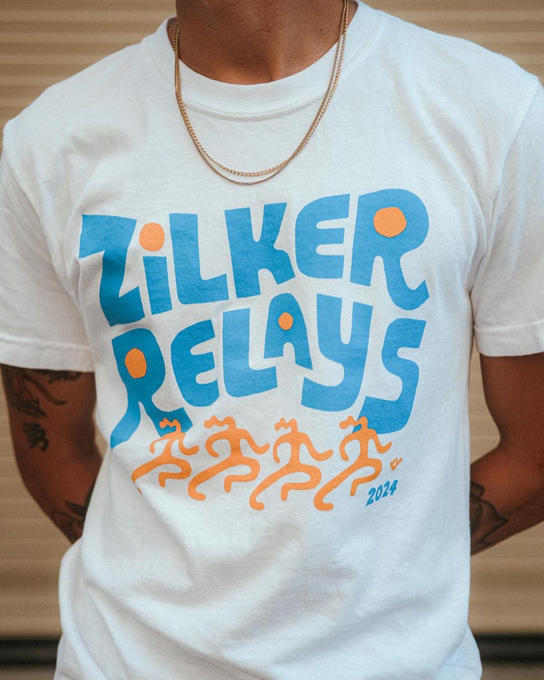 Person wearing a white t-shirt with the words 'Zilker Relays' and four figures printed on the front along with a gold chain necklace.