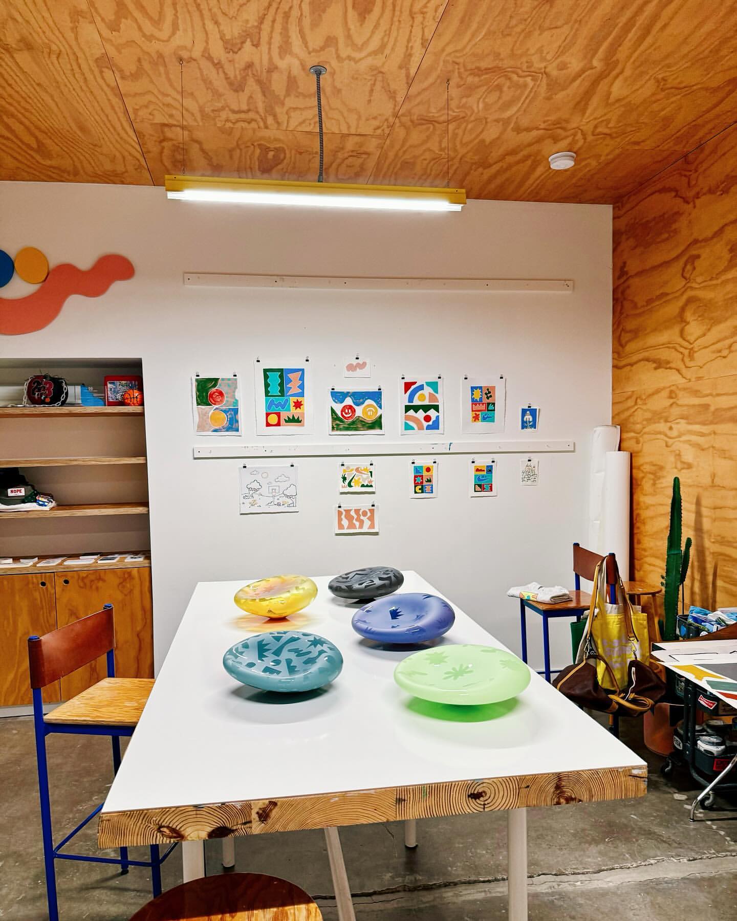 Studio space featuring a white table with colorful decorative plates, surrounded by chairs and artwork displayed on the walls.