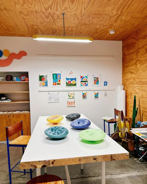 Studio space featuring a white table with colorful decorative plates, surrounded by chairs and artwork displayed on the walls.