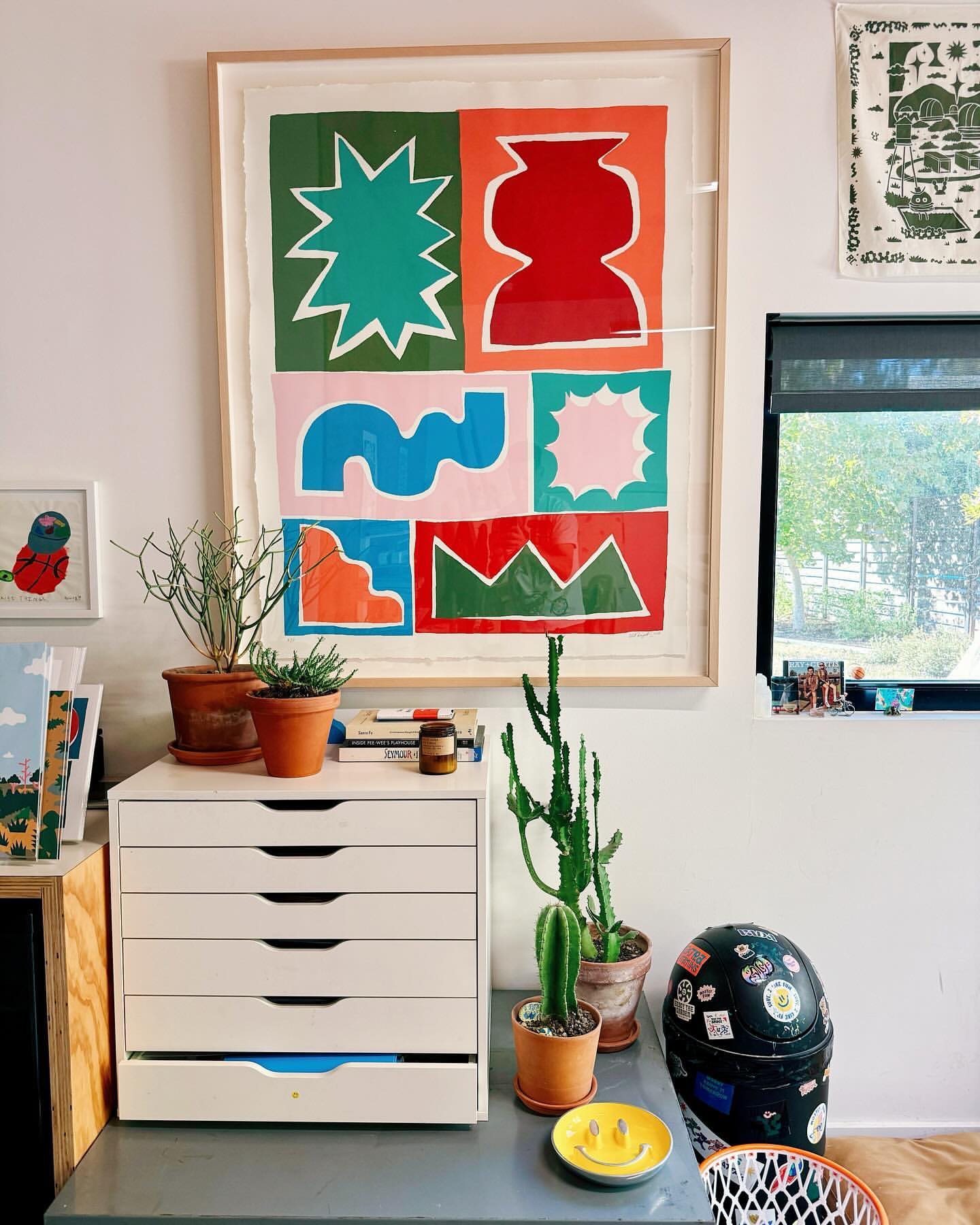 Colorful geometric abstract art in a frame hangs above a white drawer unit with potted plants in an indoor setting with natural light.