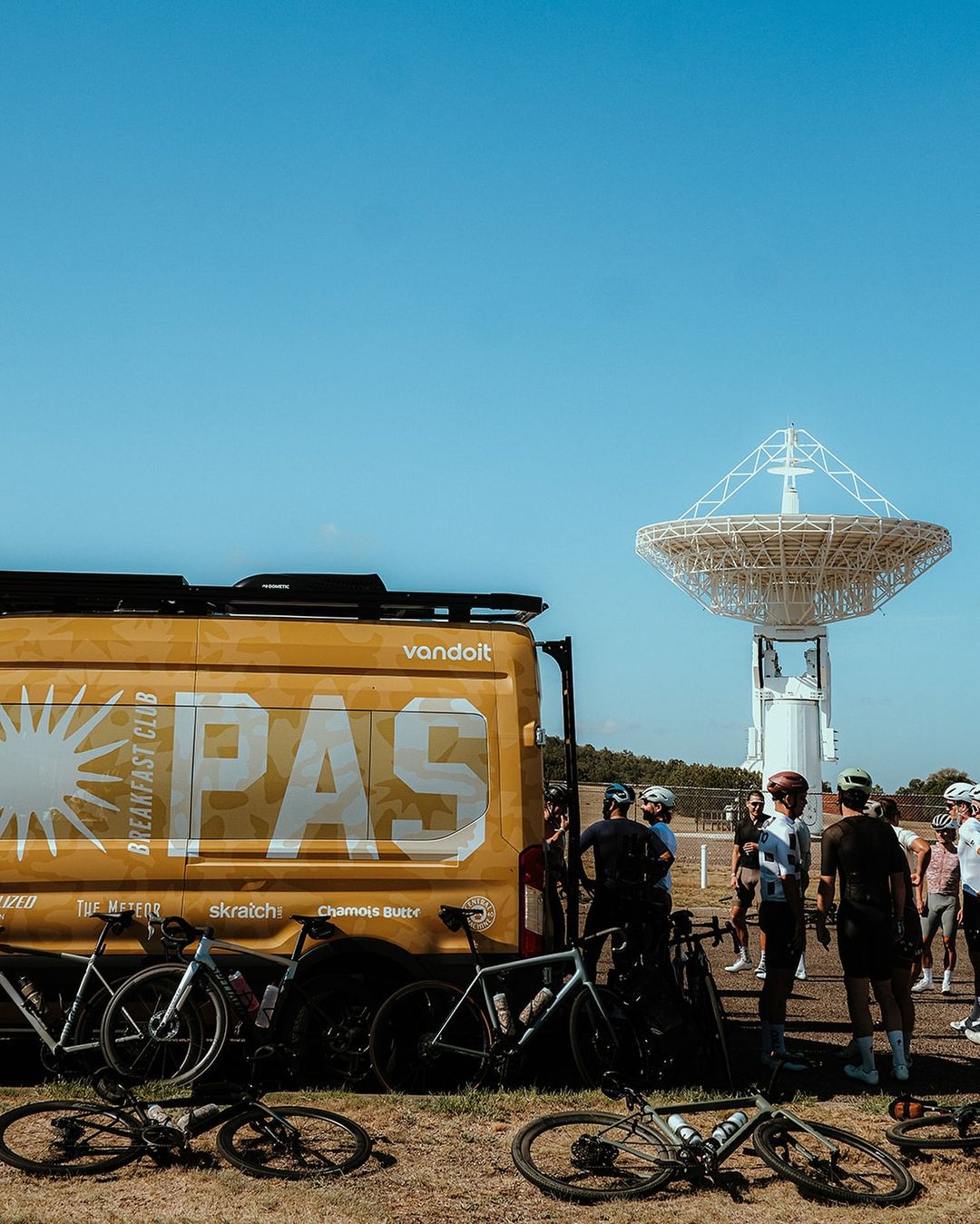 A group of cyclists gather near a van marked 'Breakfast Club PAS' and road bikes, with a large radio telescope dish in the background.