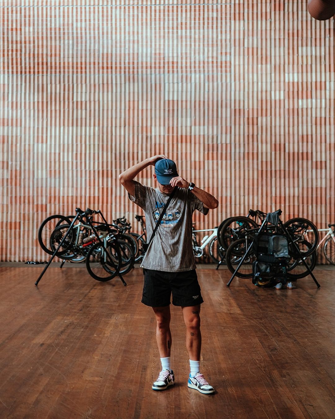 Adult in t-shirt and shorts standing in front of bikes and a striped orange wall, looking at their watch or adjusting their cap.
