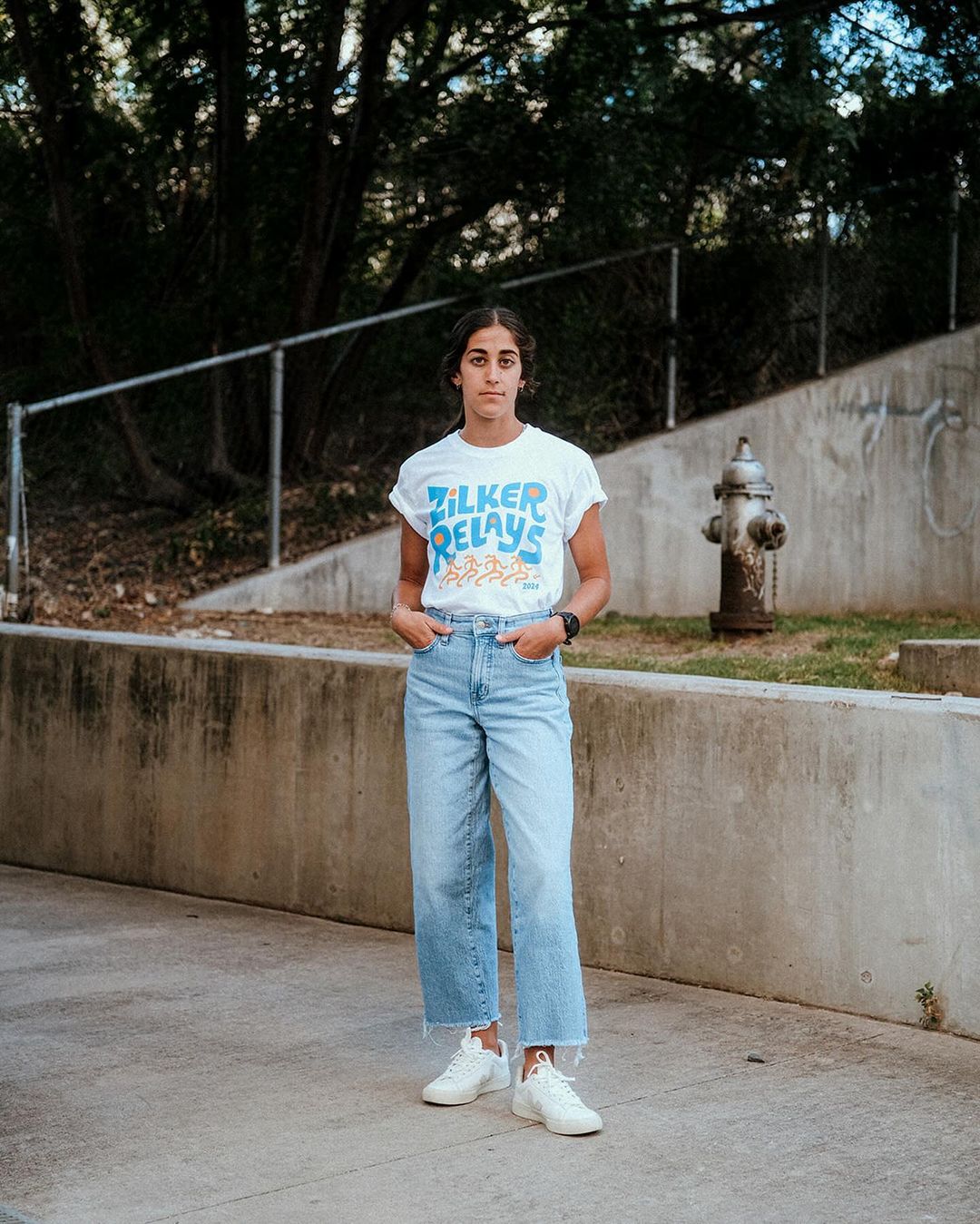 A young woman in casual clothing stands on a sidewalk in front of a concrete wall, wearing a white t-shirt, light blue jeans, and white sneakers.