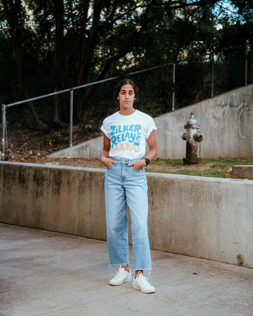 A young woman in casual clothing stands on a sidewalk in front of a concrete wall, wearing a white t-shirt, light blue jeans, and white sneakers.