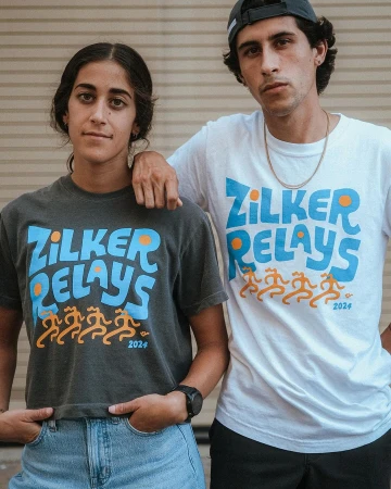 Two people wearing graphic t-shirts with Zilker Relays branding standing outdoors against a corrugated metal wall