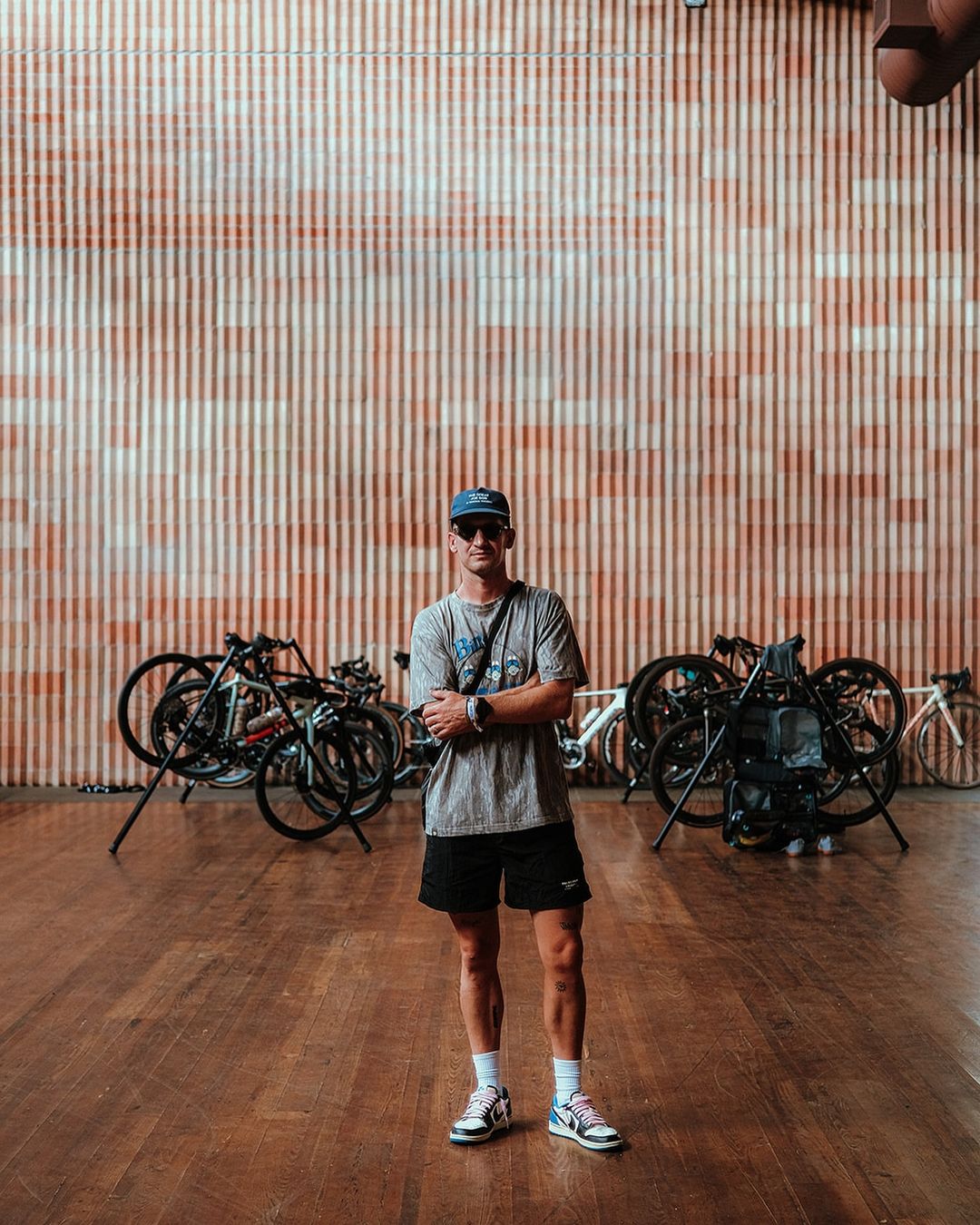Adult male stands in front of a wall and several bicycles inside an indoor space with a wood floor and natural light.