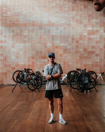 Adult male stands in front of a wall and several bicycles inside an indoor space with a wood floor and natural light.