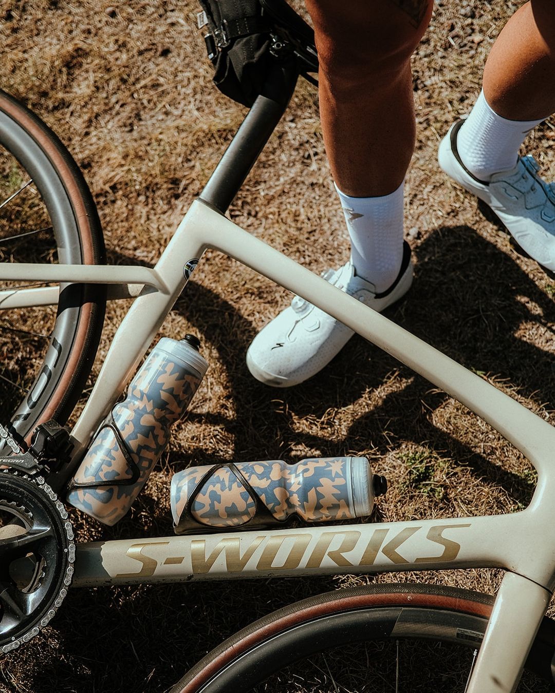 Close-up of a cream-colored S-Works bicycle lying on dry grass with two camouflage water bottles and an adult's legs and white shoes.