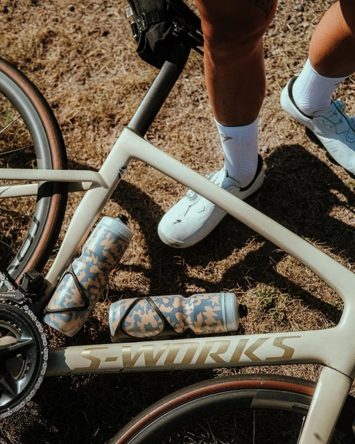 Close-up of a cream-colored S-Works bicycle lying on dry grass with two camouflage water bottles and an adult's legs and white shoes.