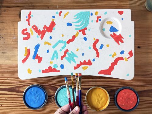 Overhead view of a white paint palette with colorful shapes, paint brushes and paint cans on a wood table for an art project.