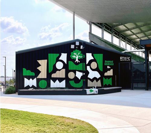 Exterior of Austin FC stadium building with a colorful mural featuring geometric shapes and a tree, accented by green grass and bright sky.