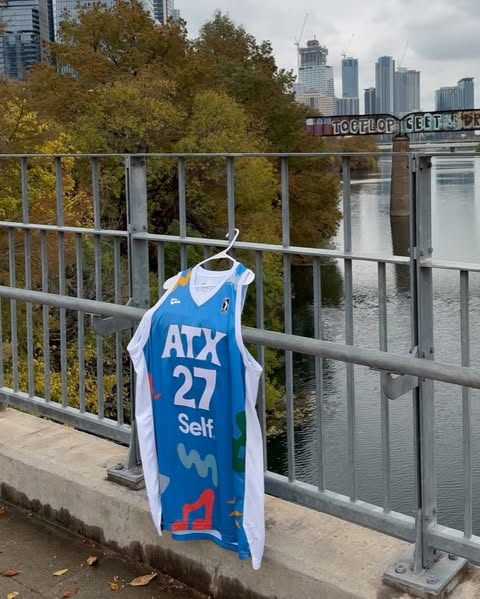 Basketball jersey hanging on a bridge railing with a city skyline and trees in the background, on a cloudy day. River below.