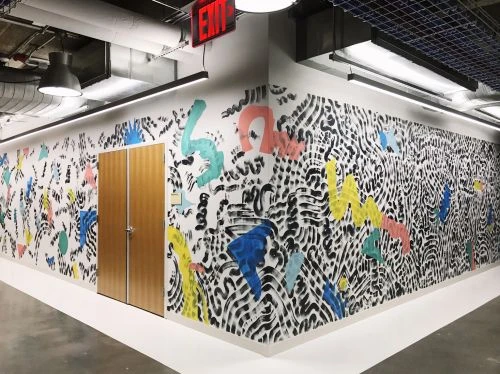 Hallway corner features large colorful graffiti-style wall art, white floors and walls, and illuminated industrial ceiling lights.
