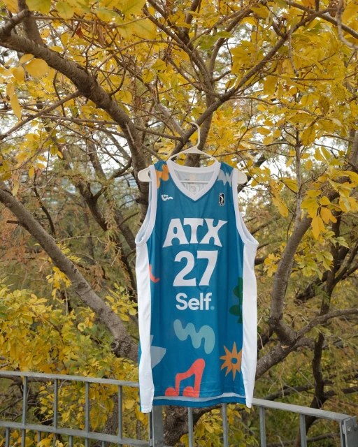Teal basketball jersey with 'ATX 27 Self' text on a hanger outdoors amid autumn trees displaying golden leaves in daylight.