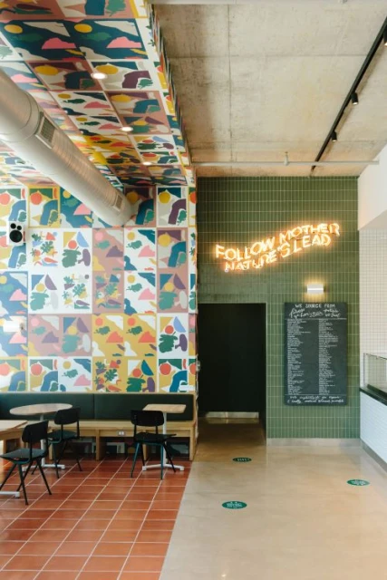 Interior of a modern cafe featuring geometric tilework, colorful abstract wall art, and neon sign reading, 'Follow Mother Nature's Lead'.