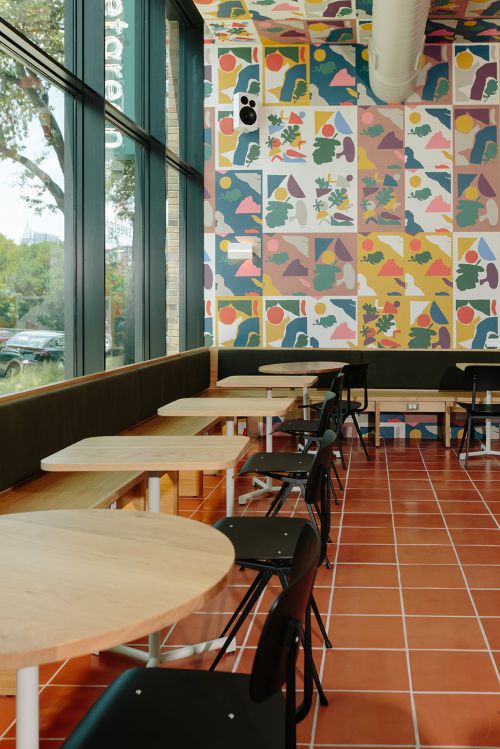 Interior of a cafe featuring wooden tables, black chairs, and colorful patterned wallpaper in natural light from large windows.