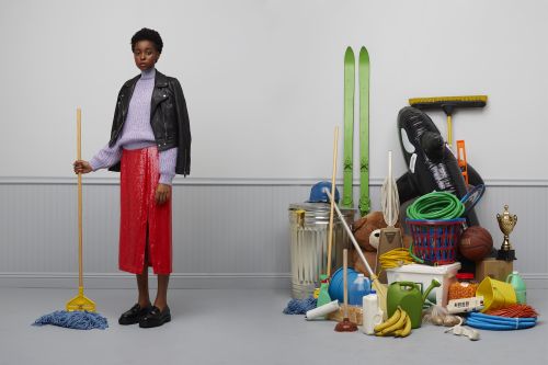 Adult female model in a red skirt and black leather jacket holds a yellow mop in an indoor studio with a pile of objects.