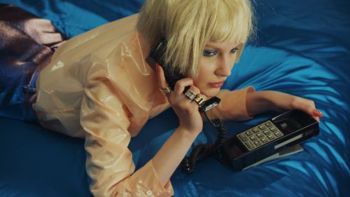 Woman in peach raincoat on blue fabric holding a black landline telephone receiver to her ear. She has short blonde hair.
