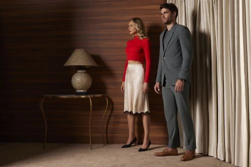 A fashionable couple poses indoors; the woman wears a red top and cream skirt, and the man wears a gray suit. Lamp and draped curtains.