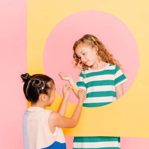 Two girls in color block outfits interact playfully through a yellow frame, with a pink backdrop. Fashionable children's wear.