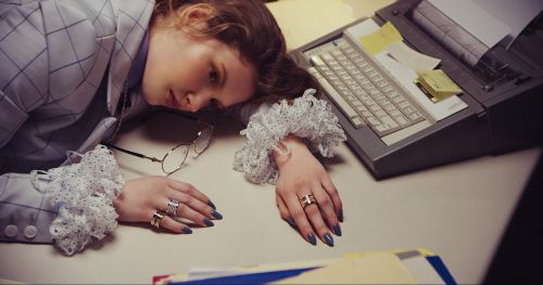 Adult woman in a checked suit lying on a desk with a typewriter, papers, and eyeglasses in an indoor setting with subdued lighting.
