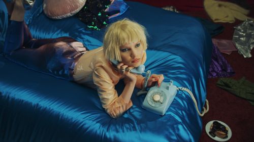 Woman lying on a blue satin bed holding a blue rotary telephone with clothes scattered around in a messy interior space.