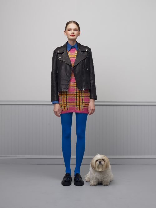 Full shot of a woman in a plaid dress and black leather jacket, standing next to a small white dog, against a grey background.