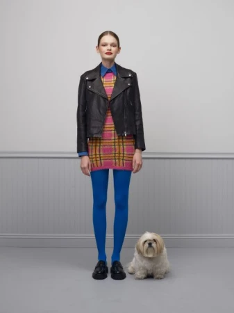Full shot of a woman in a plaid dress and black leather jacket, standing next to a small white dog, against a grey background.