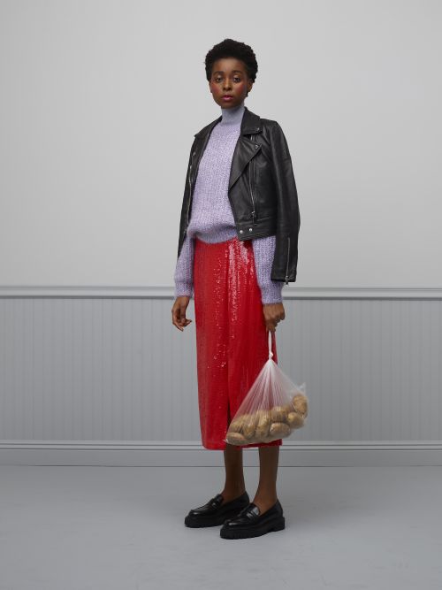 Elegant woman in a black leather jacket, lilac turtleneck, and red sequin skirt holds a bag of potatoes in a neutral indoor space.
