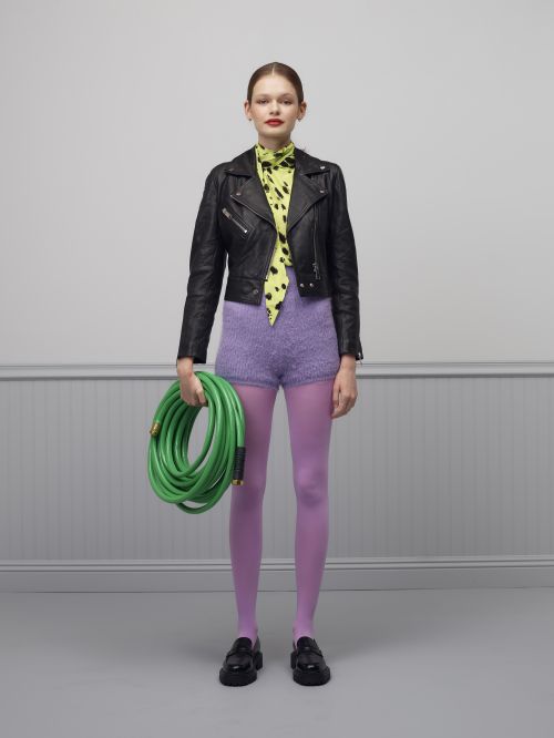Full shot of a woman in a black leather jacket and purple shorts holding a green garden hose, standing in a light gray room.