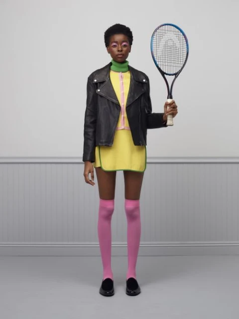 Full shot of a stylish young woman in a black jacket, yellow skirt, and pink tights, holding a tennis racket against a plain wall.