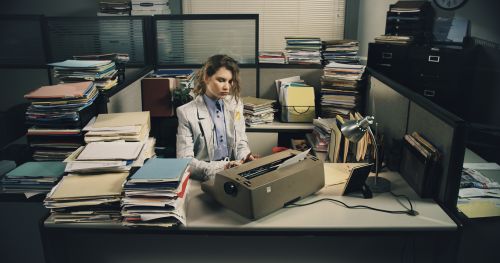 Woman works at her desk typing on a vintage typewriter surrounded by stacks of paperwork inside of an office cubicle setting.