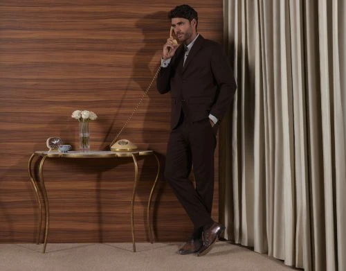 Man in a dark brown suit talking on a vintage rotary telephone against a wood panel wall with gold console table.