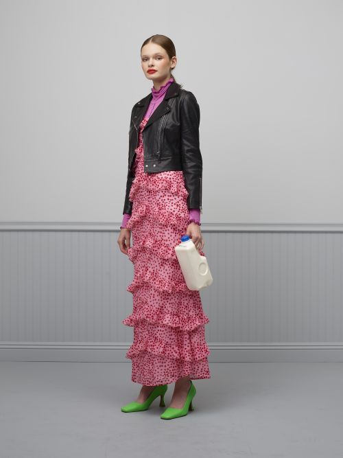Full shot of a woman in a pink dress and black jacket holding a milk jug in a gray studio with a painted wainscot.