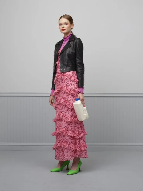 Full shot of a woman in a pink dress and black jacket holding a milk jug in a gray studio with a painted wainscot.