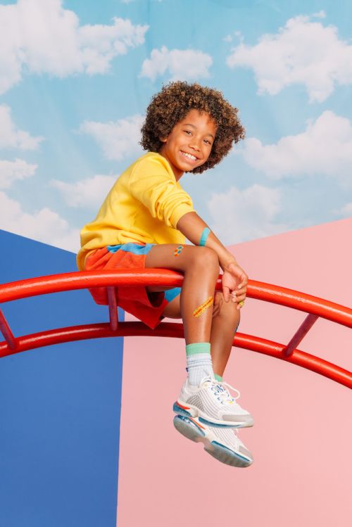 A smiling child with curly hair sits on a red play structure against a colorful sky and geometric backdrop, wearing a yellow top and shorts.