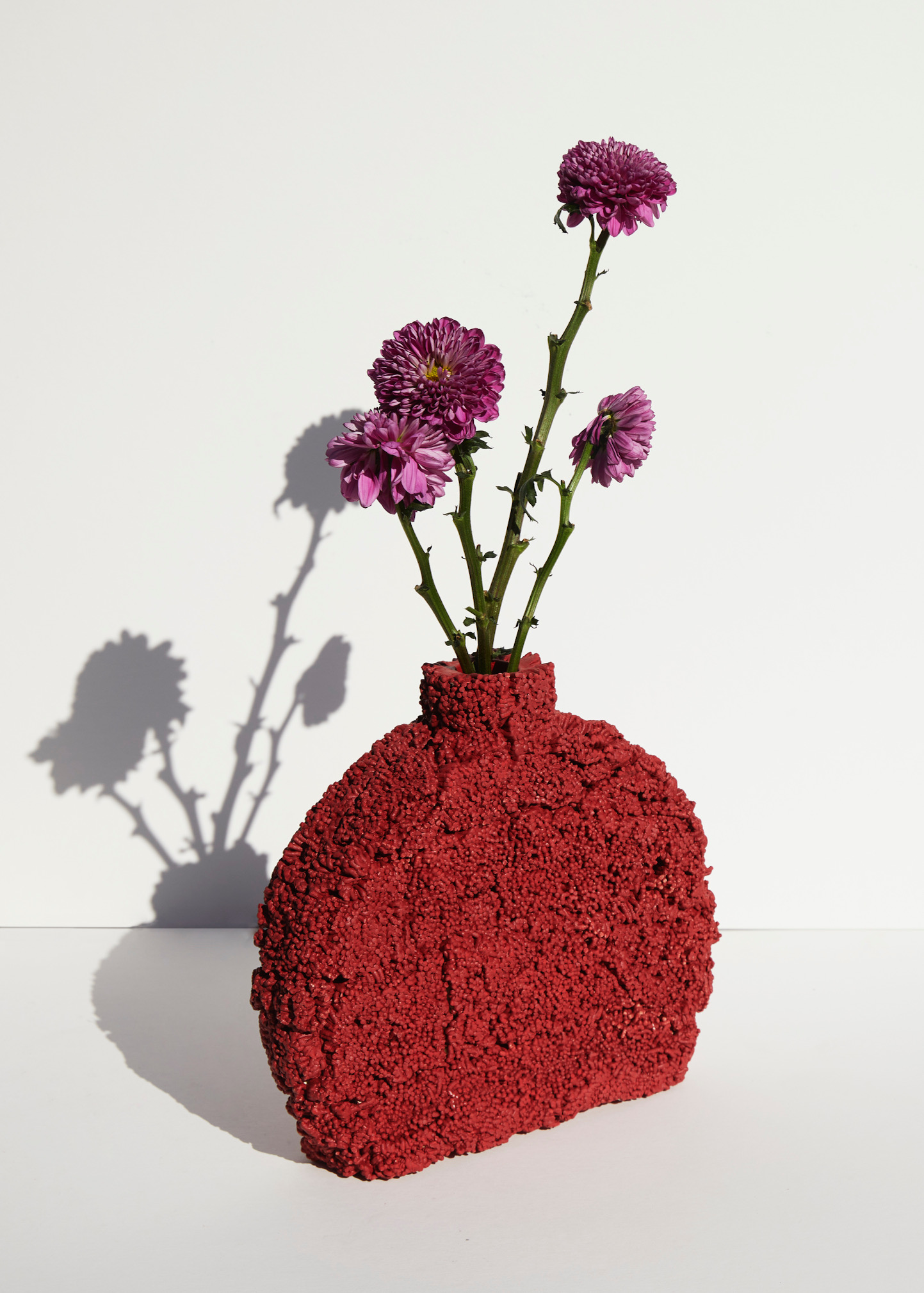 Textured red vase holding several purple mum flowers with green stems on a white surface, casting soft shadows in a studio setting.