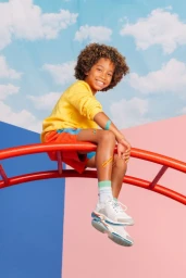A young boy with curly hair smiling while sitting on a red climbing frame against a sky and geometric pink and blue background.