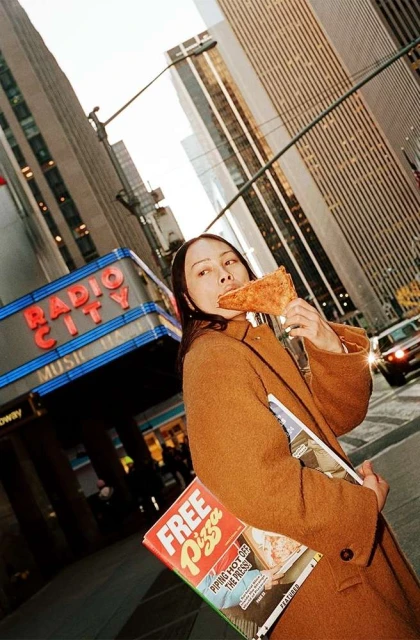 Woman in a brown coat eats pizza on a city street with skyscrapers and Radio City Music Hall visible in the background.