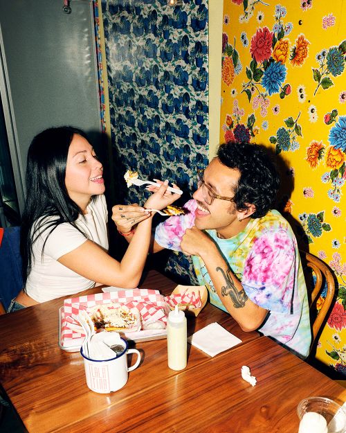 Couple smiles while feeding each other at a wooden table in front of colorful floral wallpaper, indoor setting, and natural lighting.