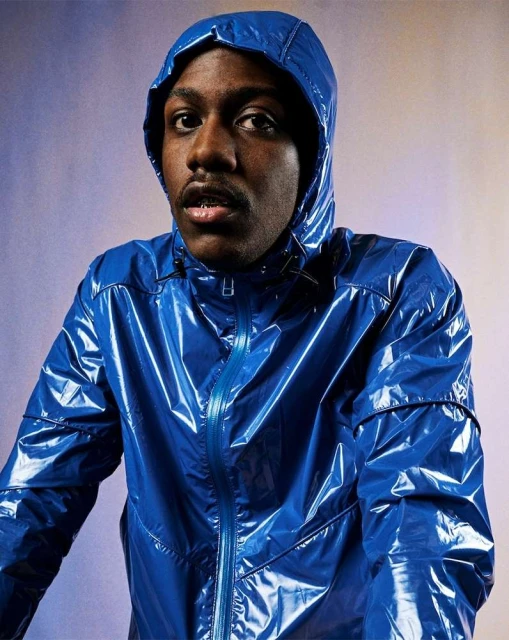 Portrait of a Black male wearing a blue hooded vinyl jacket. He looks directly into the camera with a neutral expression, indoor setting.
