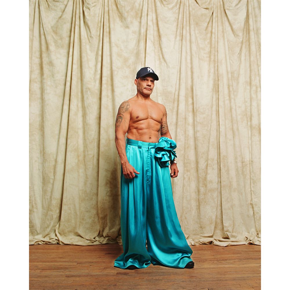 Man in stylish turquoise silk pants and baseball cap poses against a draped backdrop, showcasing fashion and fitness.