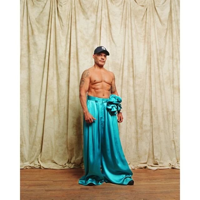 Man in stylish turquoise silk pants and baseball cap poses against a draped backdrop, showcasing fashion and fitness.