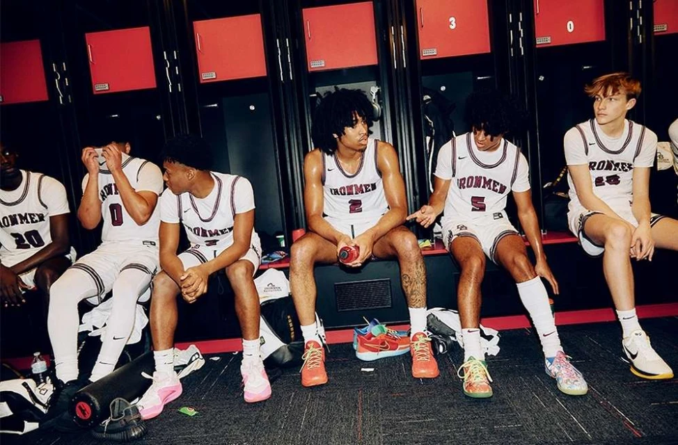 Group of six male basketball players in white uniforms with 'IRONMEN' on the front, sitting in a locker room with red lockers.