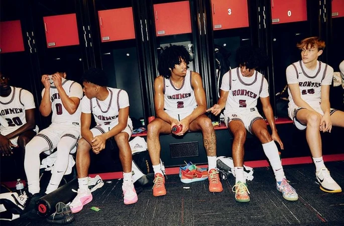 Group of six male basketball players in white uniforms with 'IRONMEN' on the front, sitting in a locker room with red lockers.